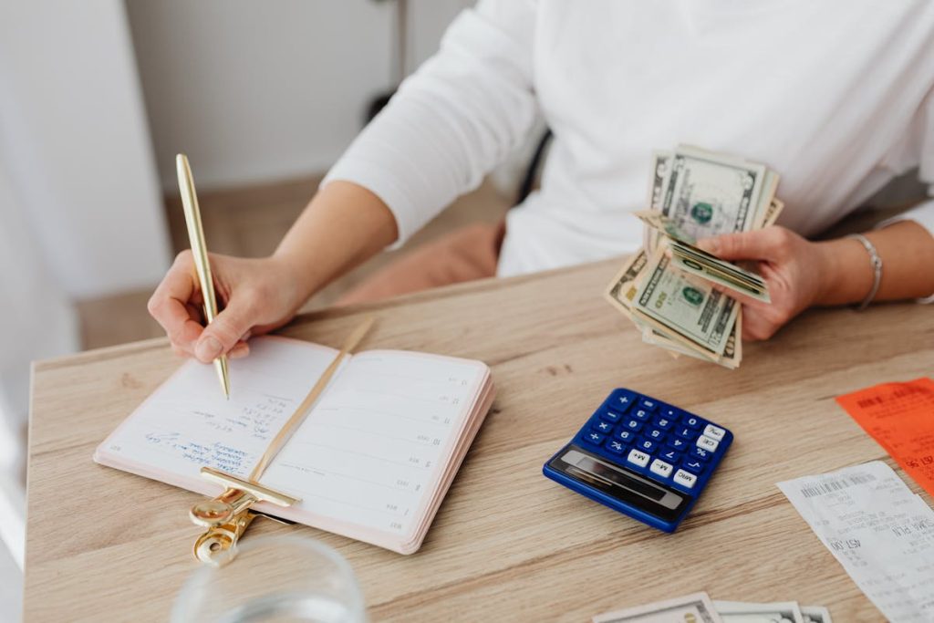 Person managing finances, writing in a planner while counting cash with a calculator and receipts on the table