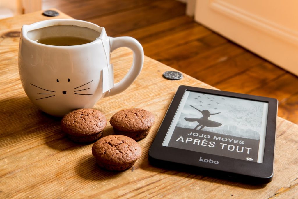 Cozy reading setup with an e-reader displaying a book, a cup of tea in a cat-themed mug, and chocolate muffins on a wooden table