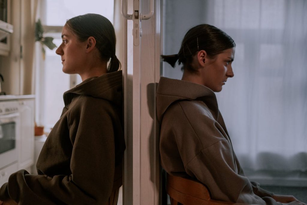 Two women sitting back-to-back against a wall, looking in opposite directions somberly