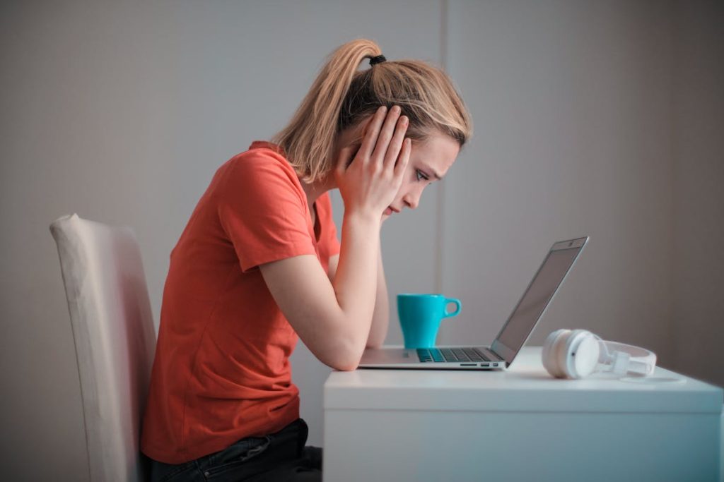 A stressed woman in a red shirt sitting at a desk, looking at her laptop with her hands on her head