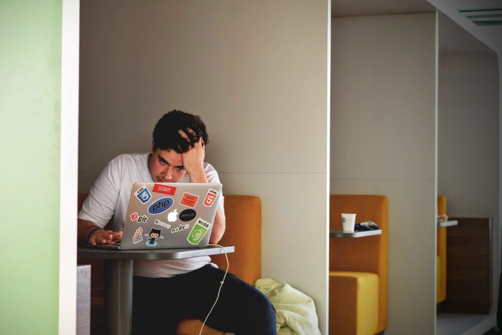 Frustrated man sitting in a booth, working on a laptop covered in tech-related stickers
