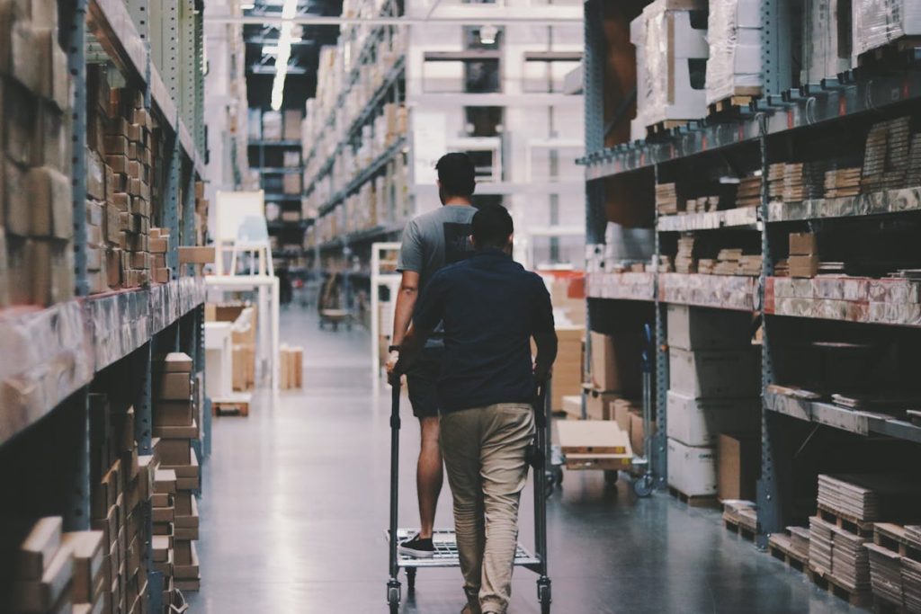 Two people pushing a cart through a large warehouse aisle filled with stocked shelves and boxes
