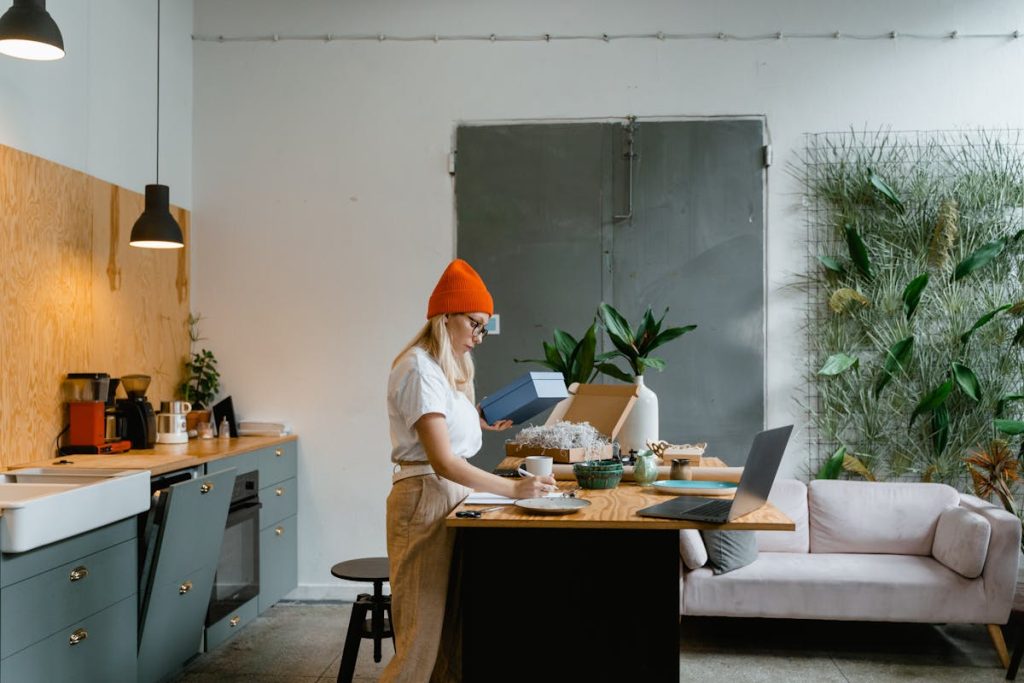 Woman wearing an orange beanie working on a laptop and packing a box in a modern, cozy kitchen