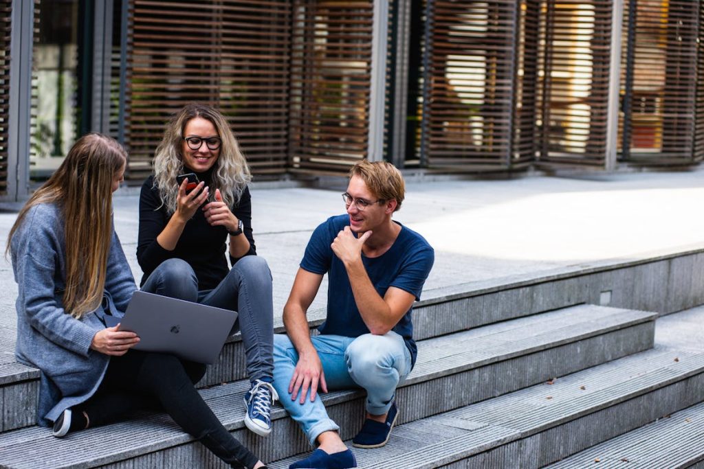 Three young adults sitting on outdoor steps, chatting and laughing. One woman is holding a phone, while another has a laptop on her lap
