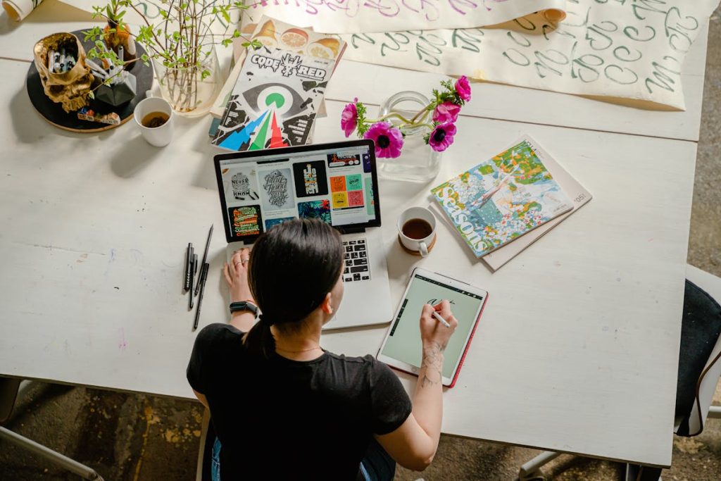 A creative designer working on a digital tablet while using a laptop, surrounded by art supplies, coffee, and magazines on a white table in a bright, artistic workspace