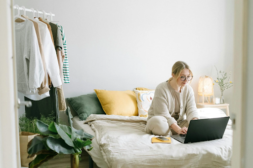 Young woman with glasses working on a laptop while sitting on a cozy bed in a minimalist bedroom with neutral tones.