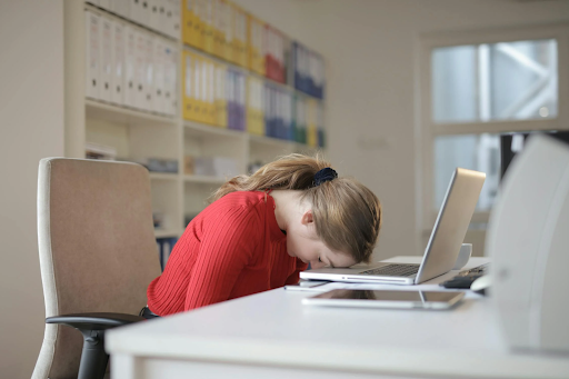 Woman in a red sweater resting her head on a desk in front of a laptop, appearing exhausted.
