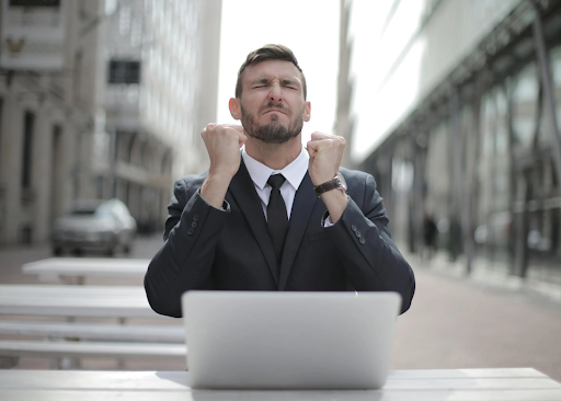 Excited businessman in a suit clenching his fists in celebration while sitting outdoors with a laptop.