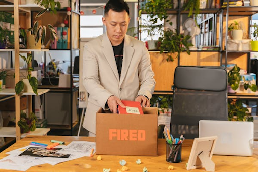 Man packing office supplies into a box labeled "FIRED" in a workspace with plants and shelves.