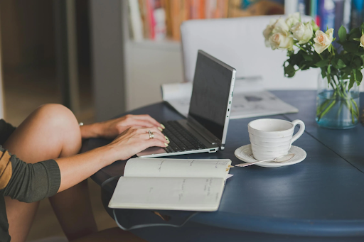 A woman working on a laptop at a cozy table with a notebook, a cup of coffee, and fresh white roses