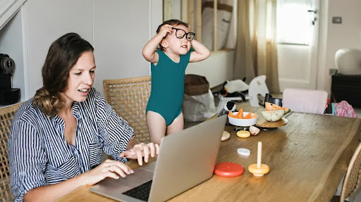 A parent working on a laptop at home, smiling with kids playing in the background.