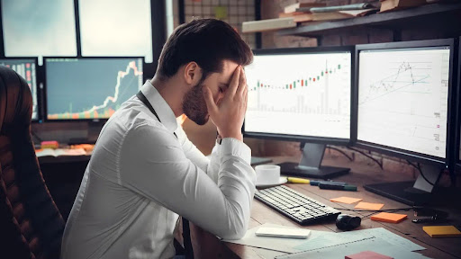 A trader sitting down with various screens towards his face as he sits in disappiontment