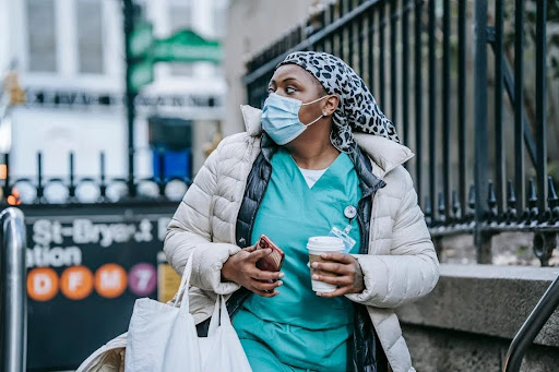 A woman in a surgical mask holds a coffee cup, joyfully walking back home