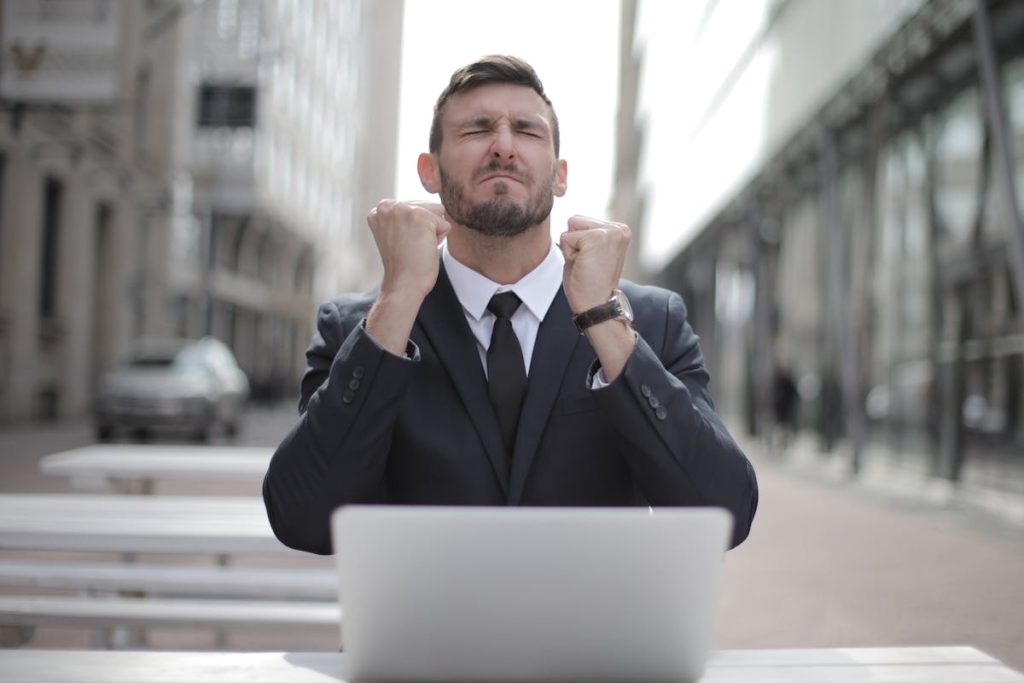 Excited businessman in a suit clenching his fists in celebration while sitting outdoors with a laptop