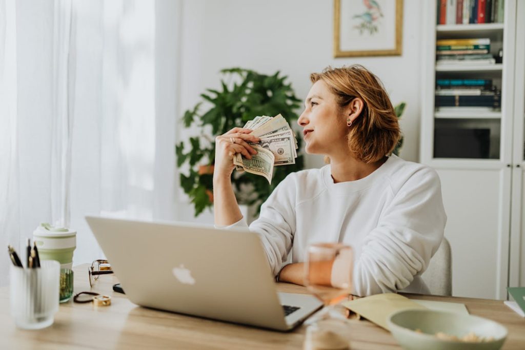 Woman holding cash beside a laptop, representing side hustle income ready to be invested