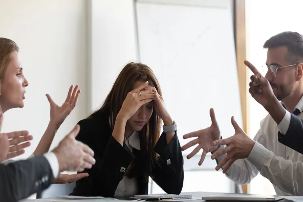 A woman with her hands on her face while surrounded by group of business professionals engaged in a meeting around a conference table